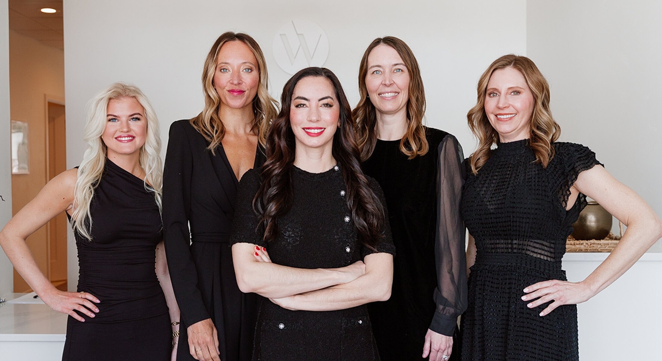 Five women in black dresses smiling together.